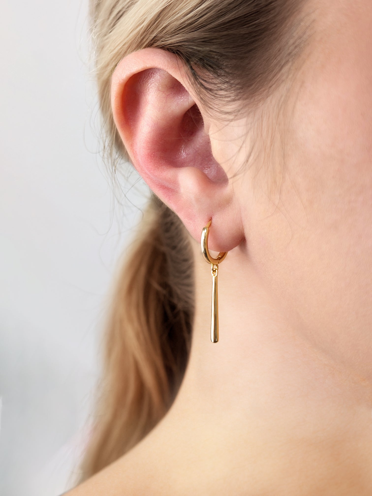 FAVES Yvette earrings in 18K gold plated sterling silver, featuring petite gold huggie hoops with slender vertical bar drops, worn on ear for a sleek and minimalist look.